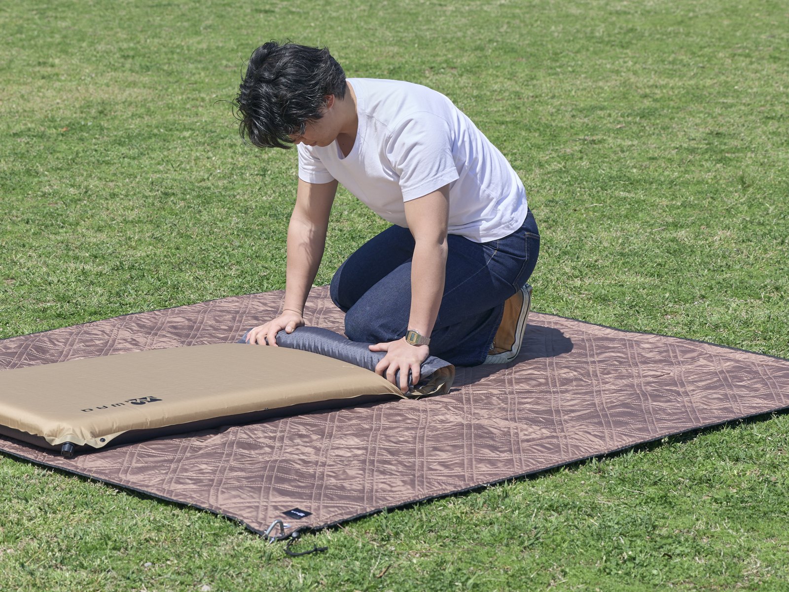 芝生に敷いたシートの上で膝をつき、インフレーターマットを端から丸めている人