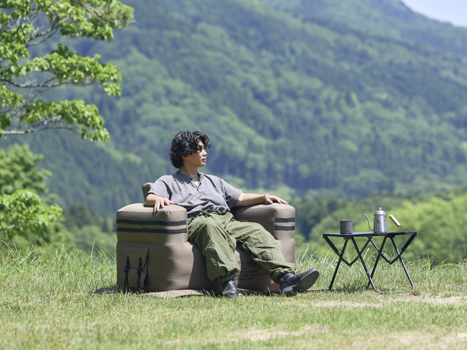山を望む草地で、コヨーテ色のシングルサイズのエアソファに1人で座る男性。横の小さなテーブルにケトルとマグ
